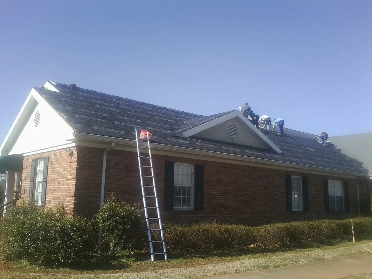 Crew tearing shingles off a brick ranch roof with a ladder set — Emergency Roof Repair work by our crew in Indian Trail