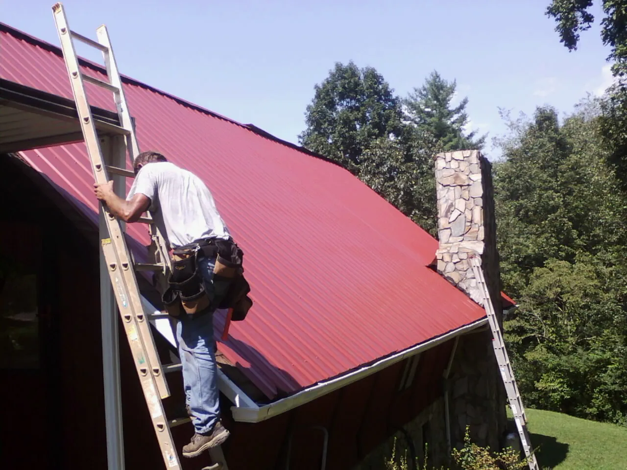 Roofer on a ladder beside a red metal roof and stone chimney — Soffit & Fascia Repair by Roofing Brothers in Indian Trail